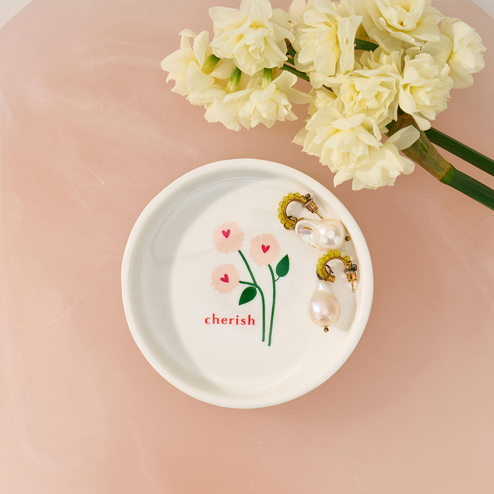 White dish with floral design and 'cherish' text, pearl earrings, and white flowers on a pink background