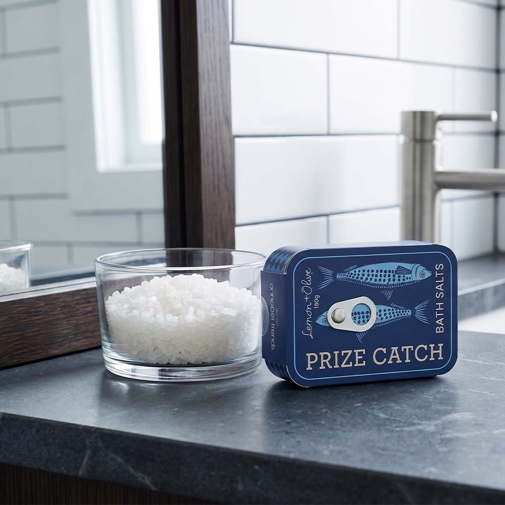 Bath salts in a glass bowl with a blue packaging box labeled 'Prize Catch' on a bathroom counter.