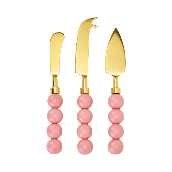 Three gold cheese knives with pink bead handles on a white background