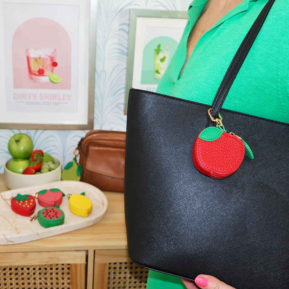 Black bag with a red apple charm held by a person in a green shirt, with fruit-themed decor in the background.