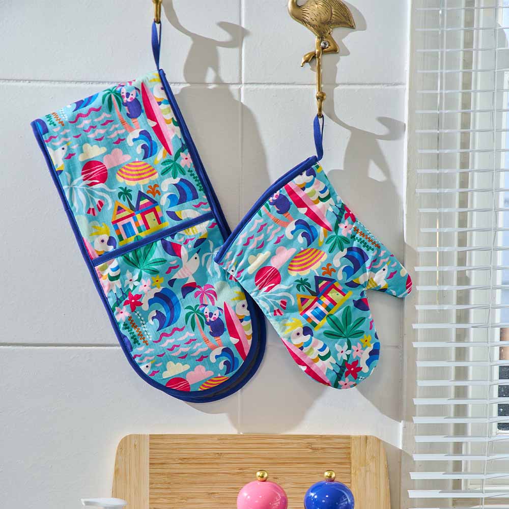 Colorful oven mitts with a tropical pattern hanging on a hook against a white tiled wall.