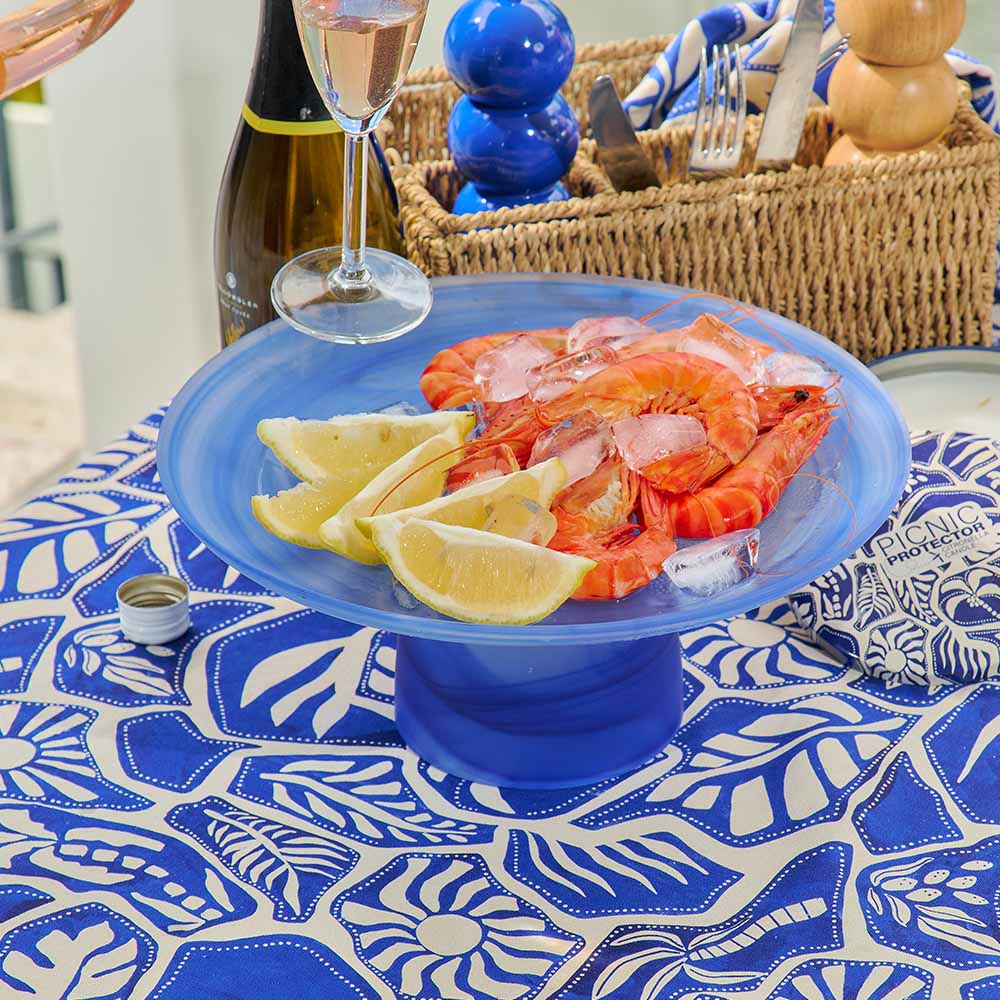 Platter of shrimp with lemon wedges on a blue stand, set on a tablecloth with a blue and white pattern.
