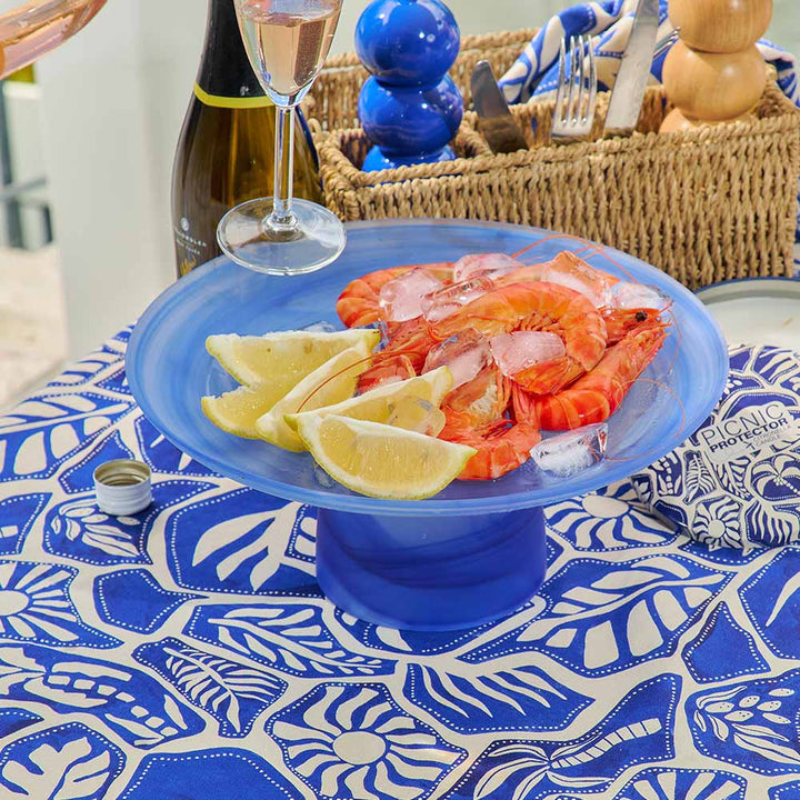 Platter of shrimp with lemon wedges on a blue stand, set on a tablecloth with a blue and white pattern.