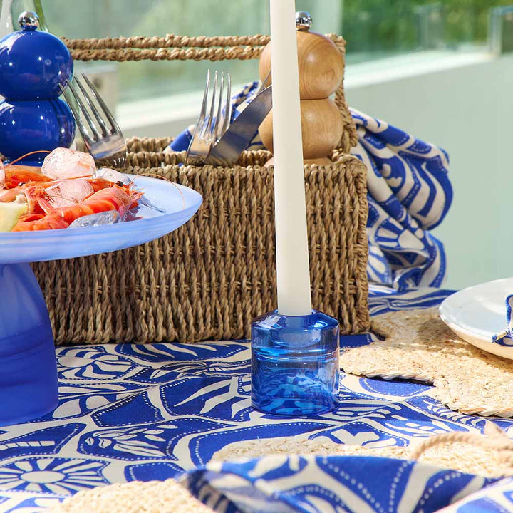 Table setting with blue and white patterned tablecloth, wicker basket, and blue candle holder.