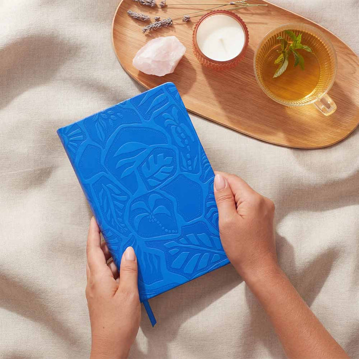 Blue notebook with embossed design held by hands on a light surface with a wooden tray featuring a cup of tea, candle, and rocks.