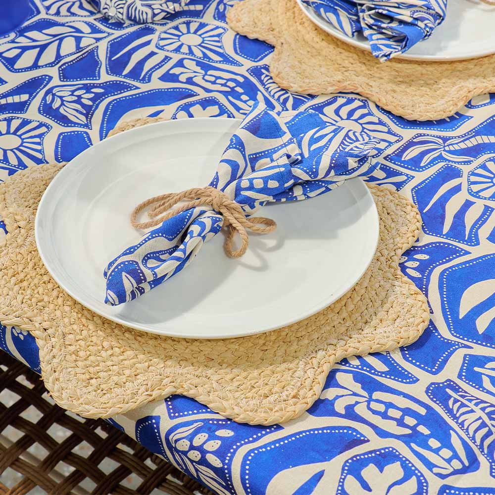 Table setting with white plates, blue patterned tablecloth, and woven placemats.