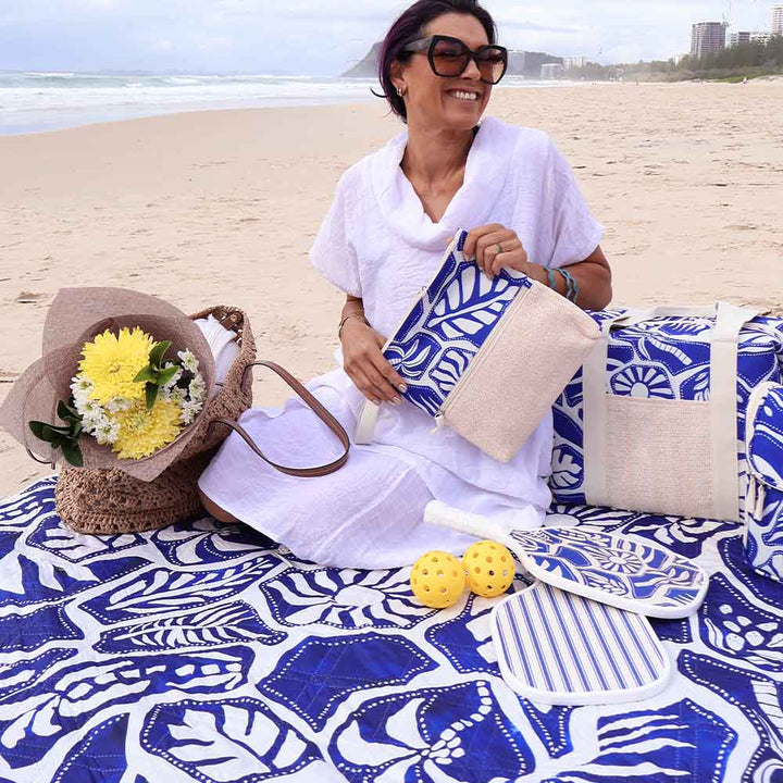 Woman sitting on a beach with a blue and white patterned blanket, holding a bag with flowers.