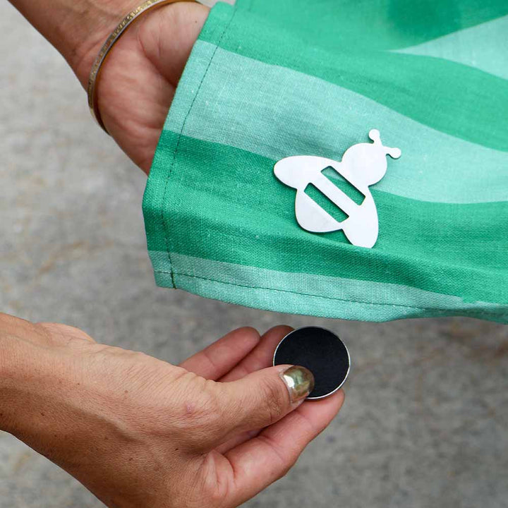 Green fabric with a white bee tablecloth weight held by a person, with a small black object on a gray surface.