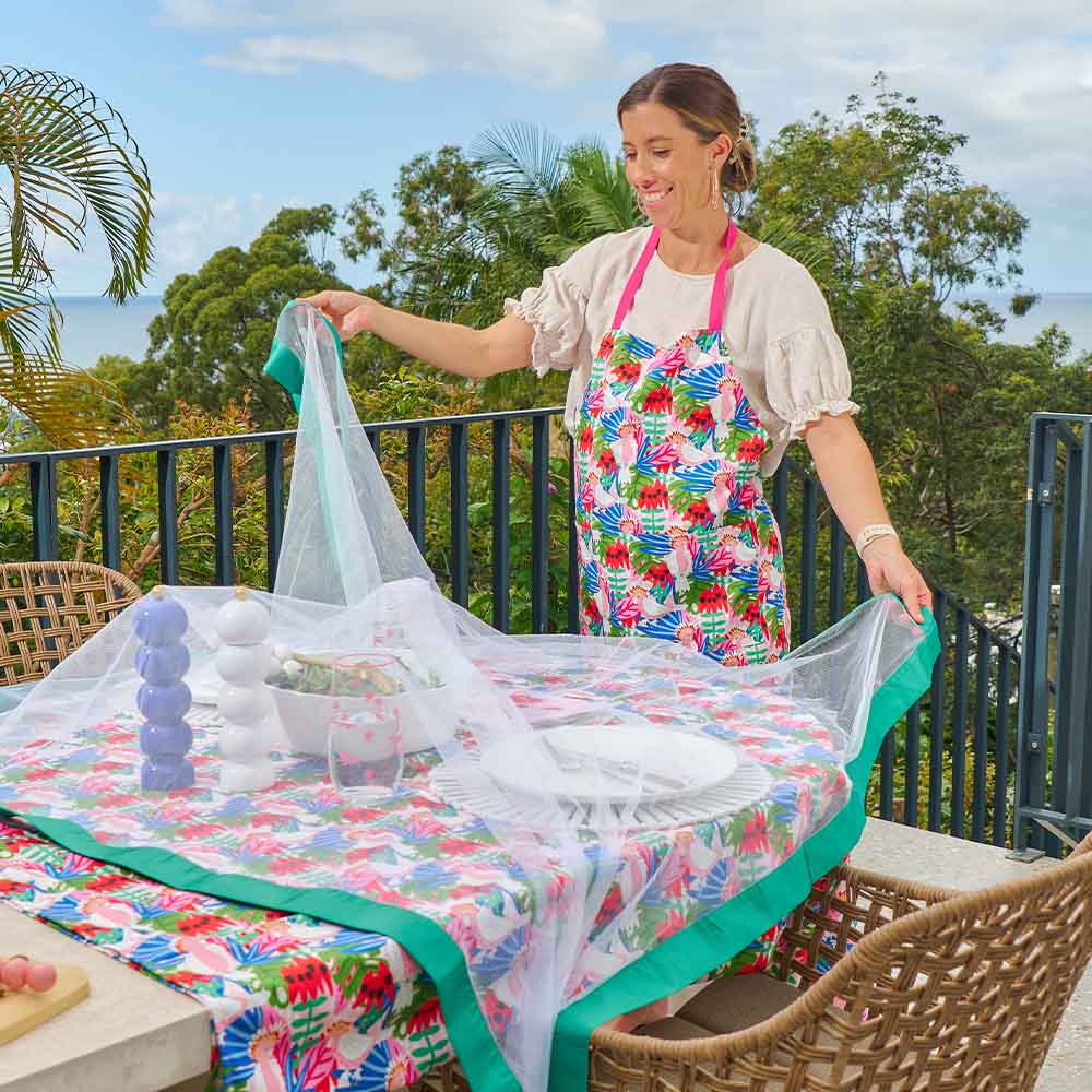 Woman setting up a table with a floral tablecloth and apron on a balcony with trees in the background