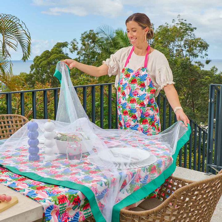 Woman setting up a table with a floral tablecloth and apron on a balcony with trees in the background