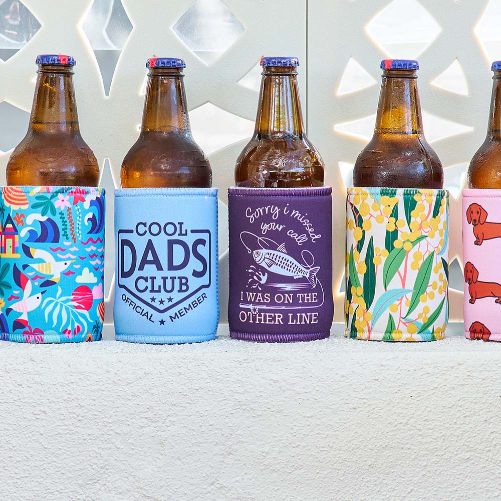 Five bottles with colourful can cooler on a textured surface with a geometric patterned wall in the background.