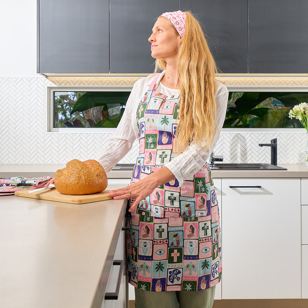 Woman in a kitchen wearing a colorful apron with a loaf of bread on a cutting board.