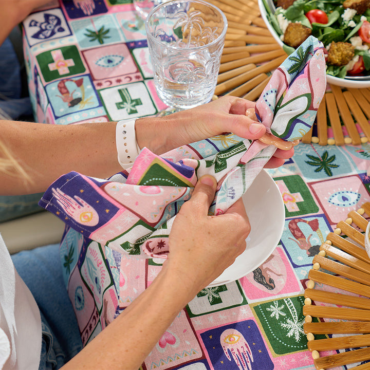 Person folding a colorful napkin on a patterned tablecloth with a glass of water and salad in the background.