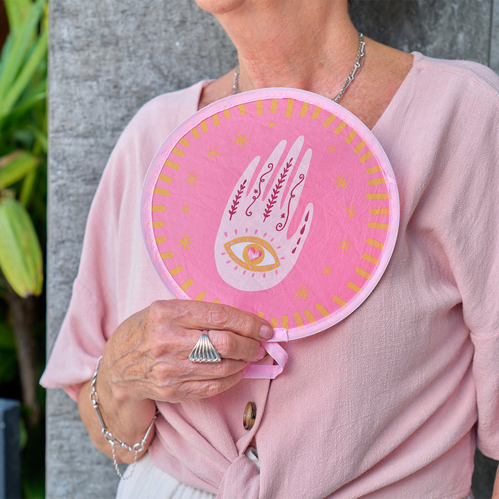 Person holding a pink hand twist fan with celestial dreams hand design against a gray wall.