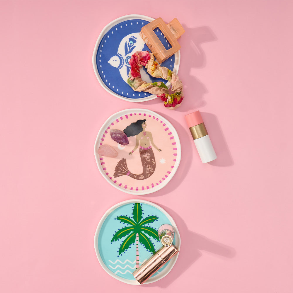 Three decorative trinket dishes with mermaid, palm tree, and fish designs on a pink background.
