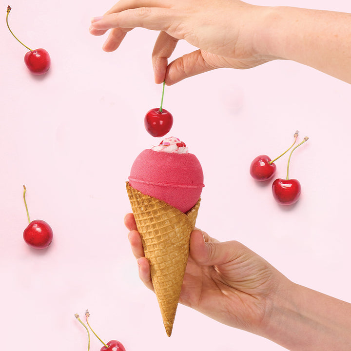 Bath bomb in a waffle cone with cherry on top held by hands against a pink background