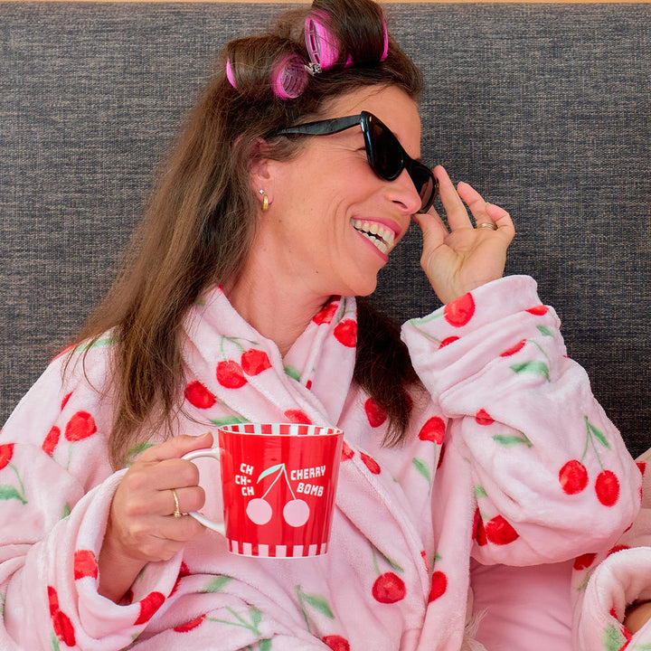 Woman in a pink robe with cherry pattern holding a red cup, wearing sunglasses and hair rollers.