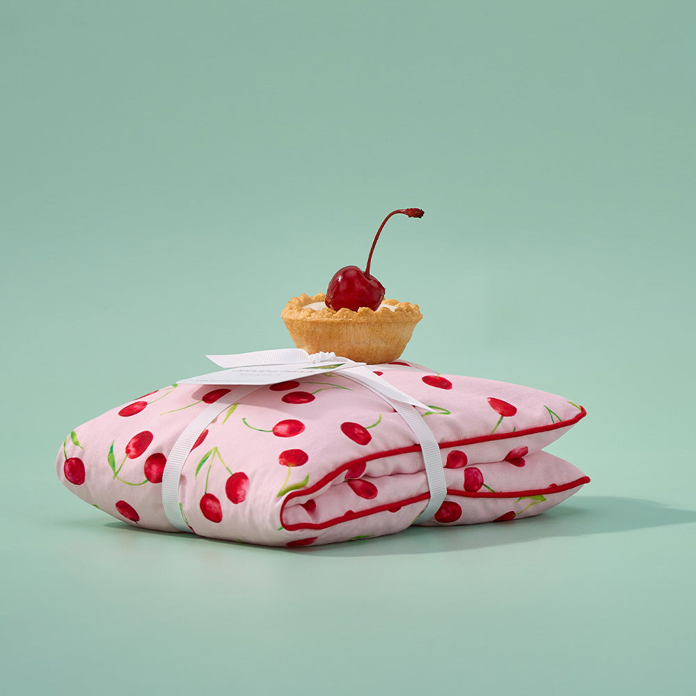 Pink heat pillow with cherry pattern and a small tart with a cherry on top against a light green background