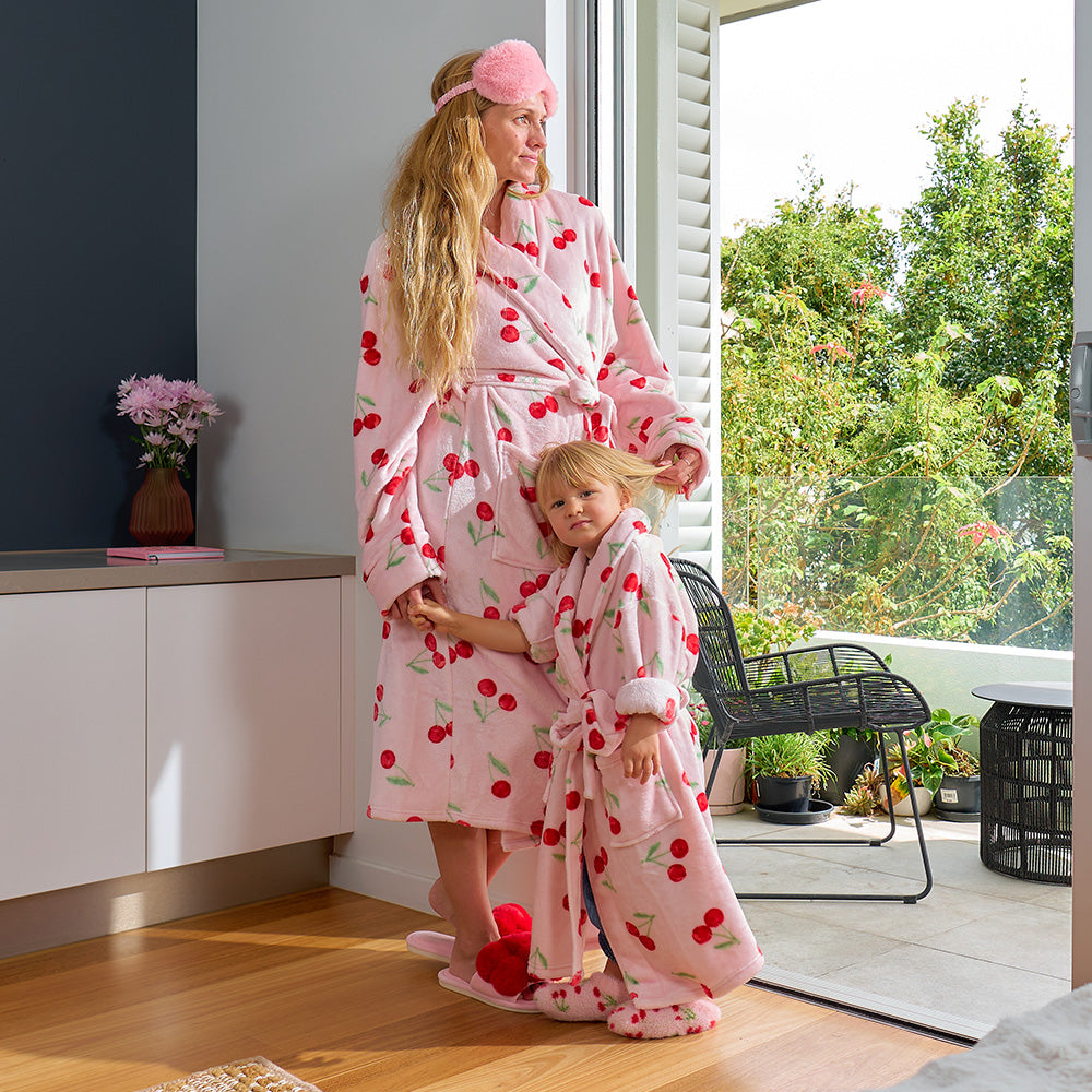 Woman and child in matching pink robes with cherry pattern standing in a room with plants and a window.
