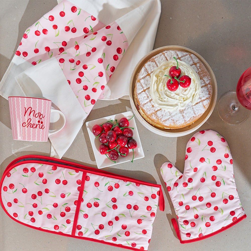 Cherry-themed kitchen items including oven mitts, pot holder, and mug on a table with a cake and cherries.