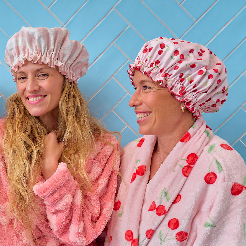 Two women wearing pink shower caps and robes against a blue tiled wall.