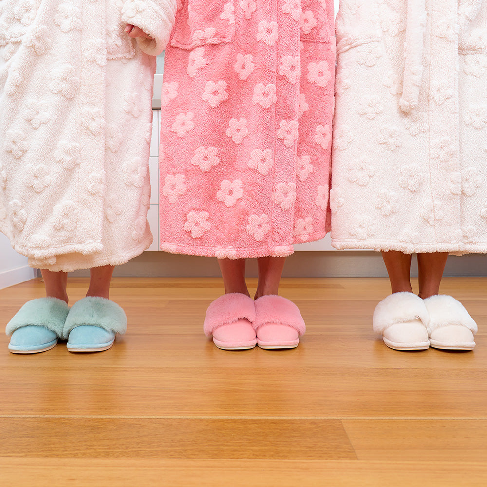 Three pairs of feet wearing colorful slippers and floral bathrobes on a wooden floor.