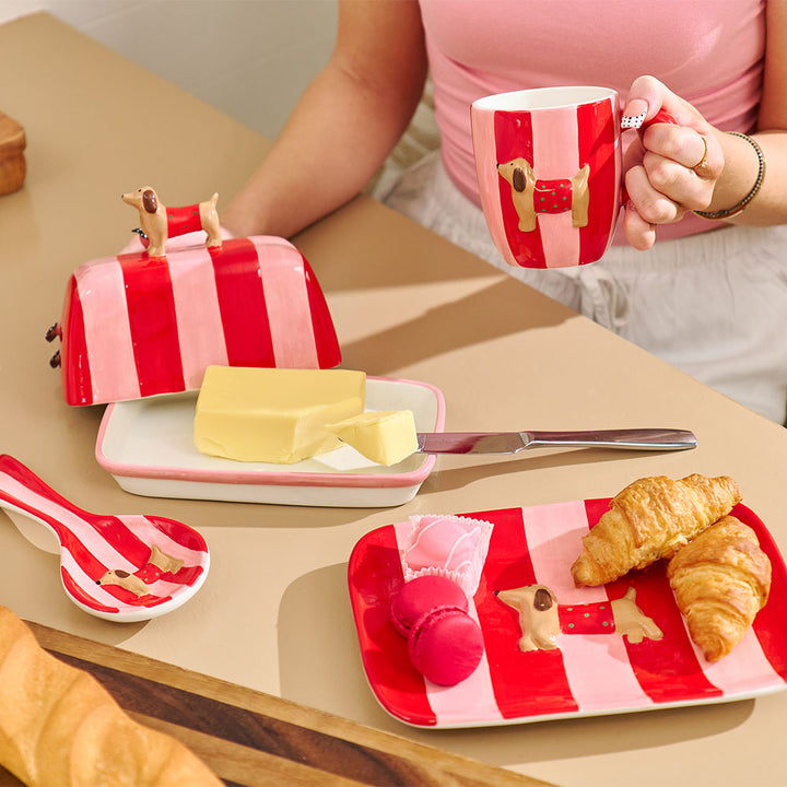 Person holding a red and white striped mug with a breakfast setup on a table.