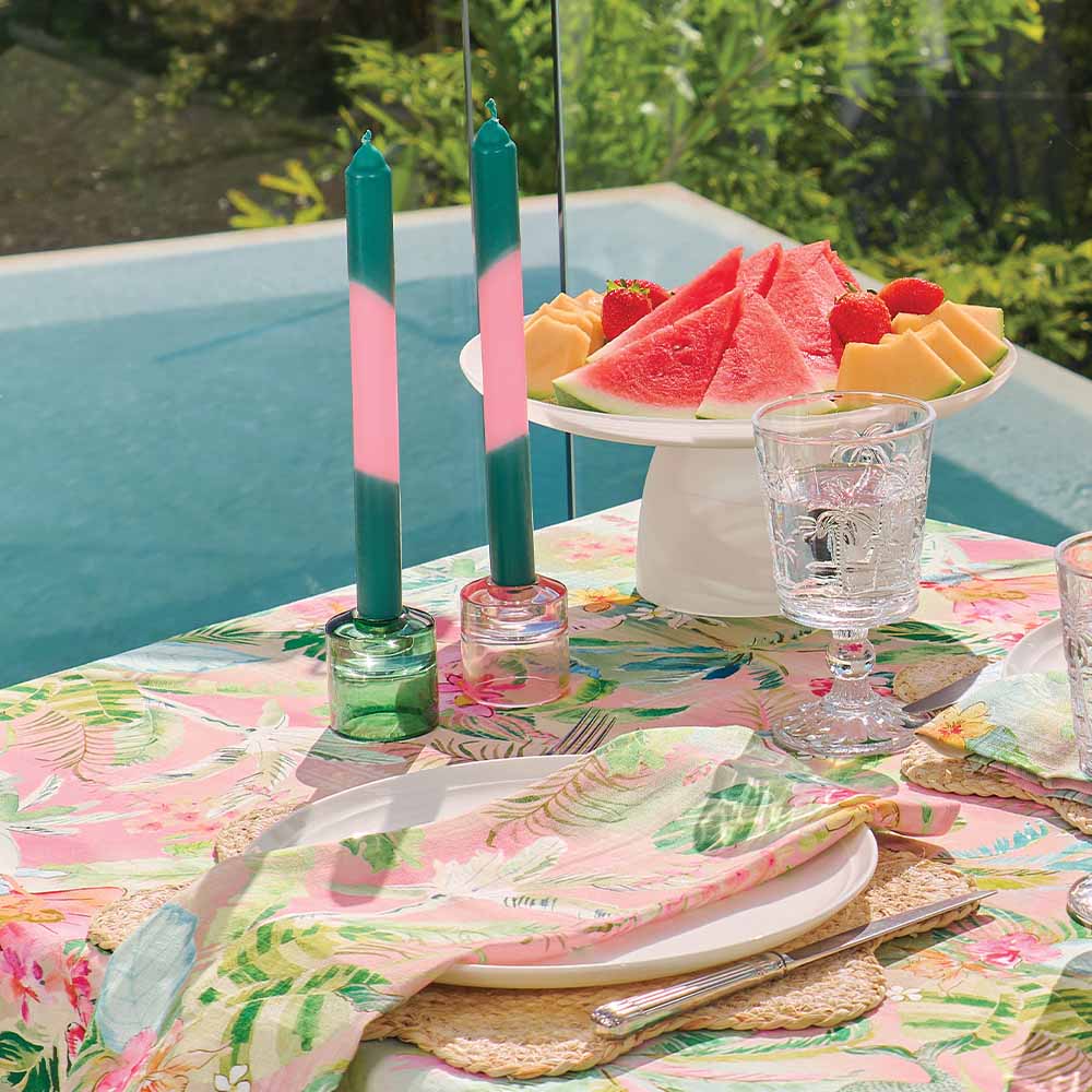 Table setting with watermelon, fruit, and candles on a floral tablecloth by a pool.