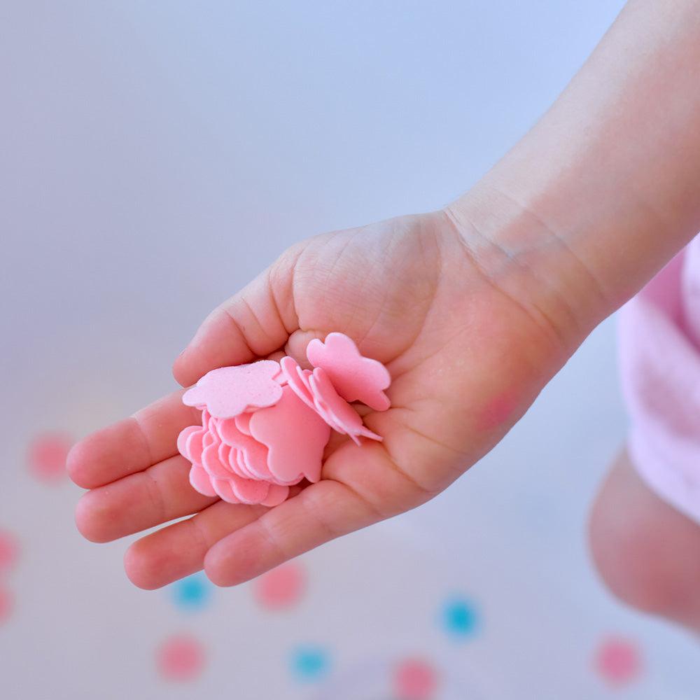 Child's hand holding a pink flower-shaped object against a blurred background with colorful dots.