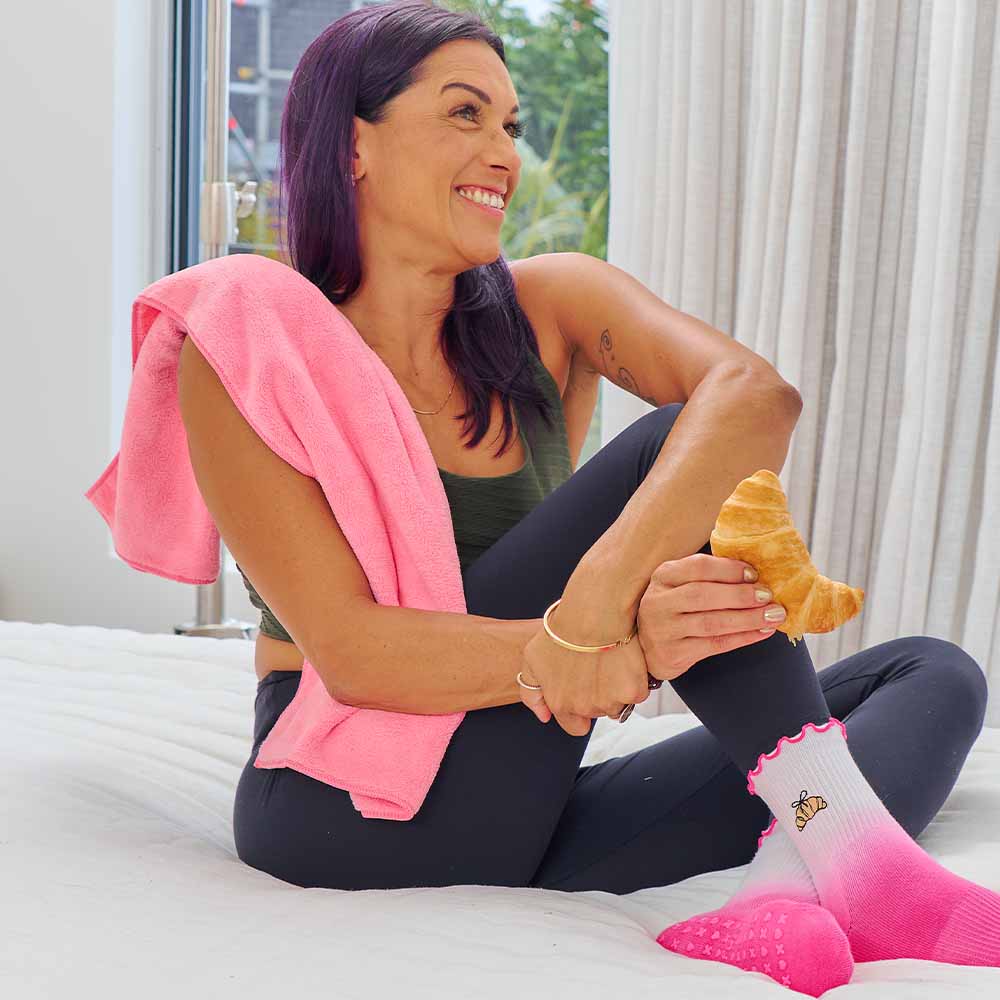 Woman sitting on a bed holding a croissant with a pink towel draped over her shoulder.