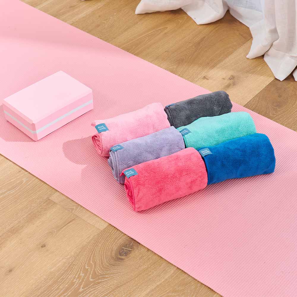 Set of colorful towels on a pink yoga mat with a block on a wooden floor.