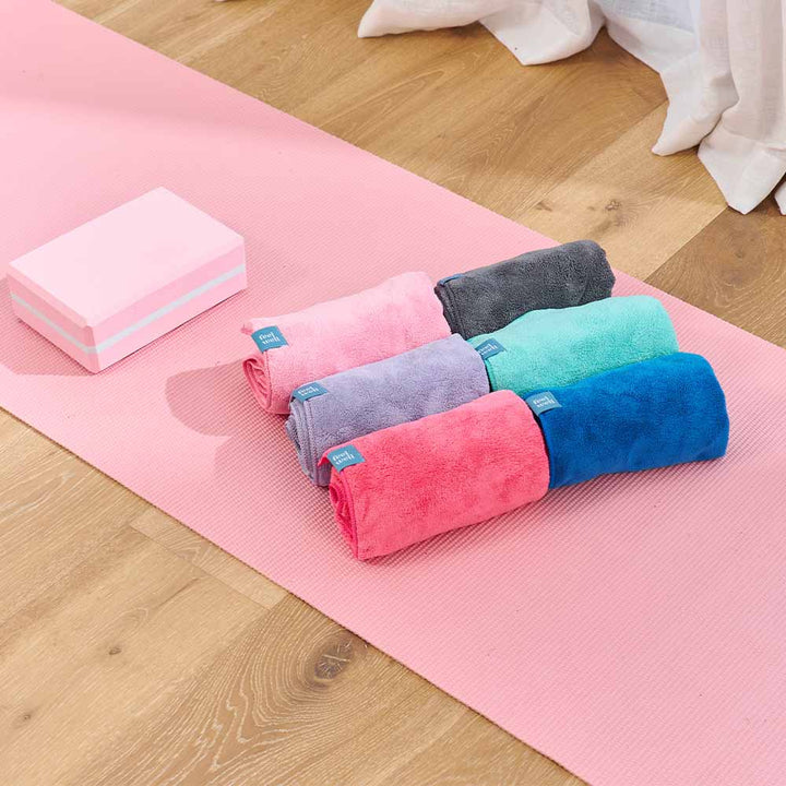 Set of colorful towels on a pink yoga mat with a block on a wooden floor.