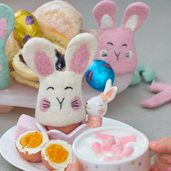 Felt bunny egg warmer with a cup of hot chocolate, Easter eggs, and pastries on a grey background.