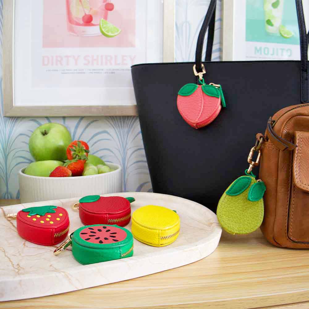 Colorful fruit-shaped keychains attached to bags on a table with a fruit bowl in the background.