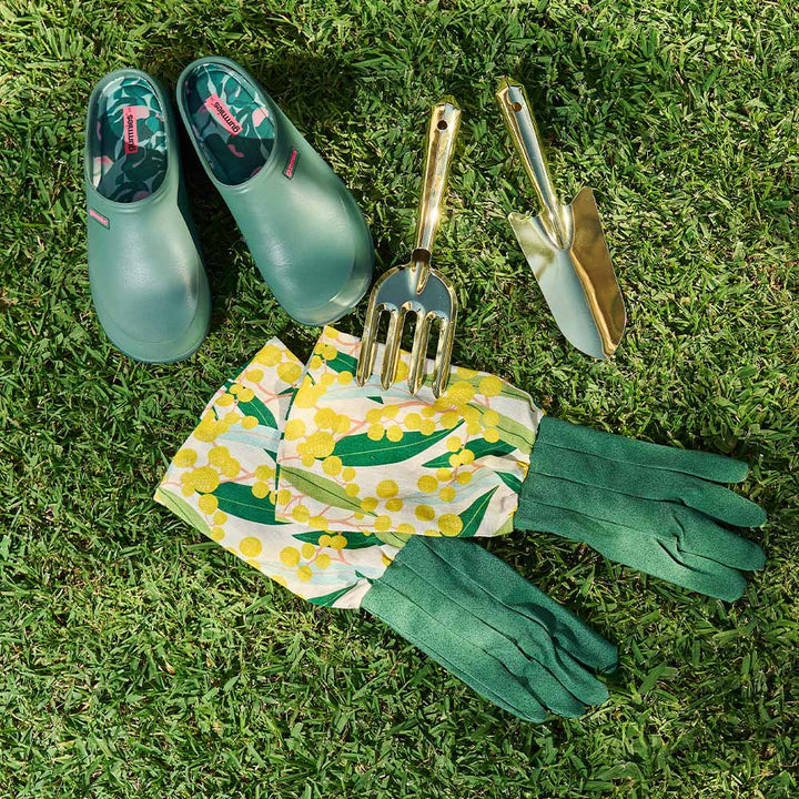 Gardening tools and accessories including green shoes, gloves, and a floral apron on grass.