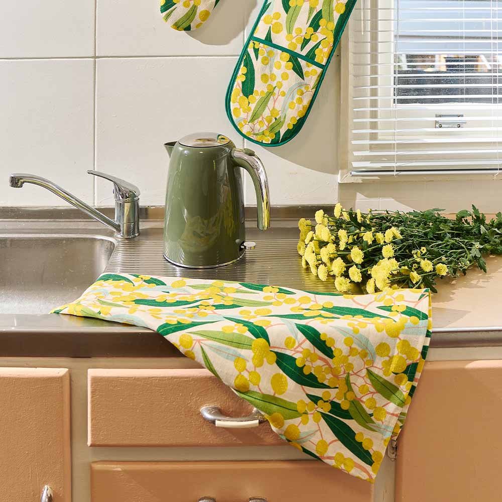 Kitchen scene with a green kettle, yellow floral towel, and flowers on a counter.