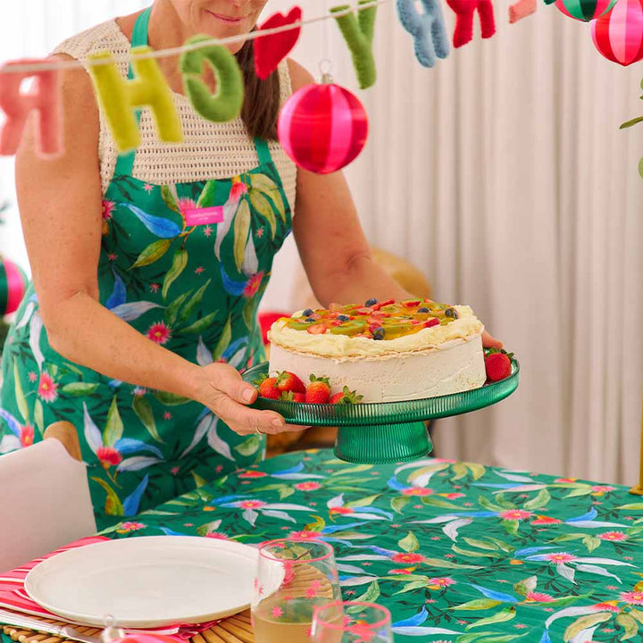 Person in a floral apron holding a cake with colorful decorations, surrounded by festive elements.