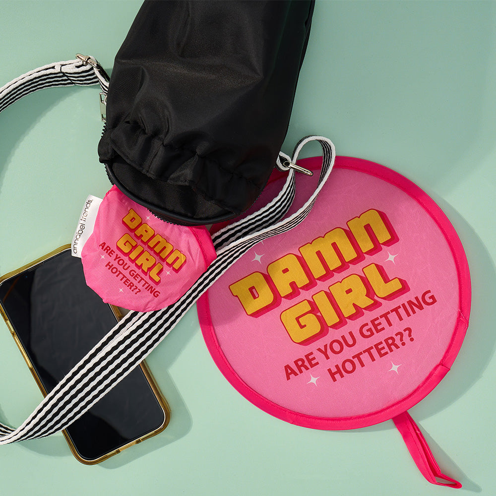 Pink pouch with 'Damn Girl Are You Getting Hotter?' text, black jacket, and phone on a light green background