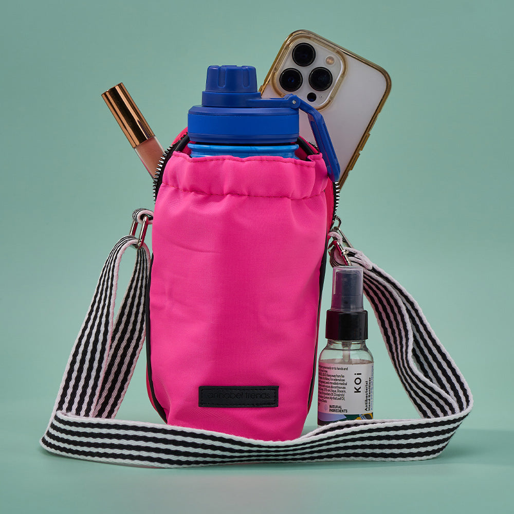 Pink crossbody bottle bag with striped strap containing a phone, water bottle, and other items on a green background