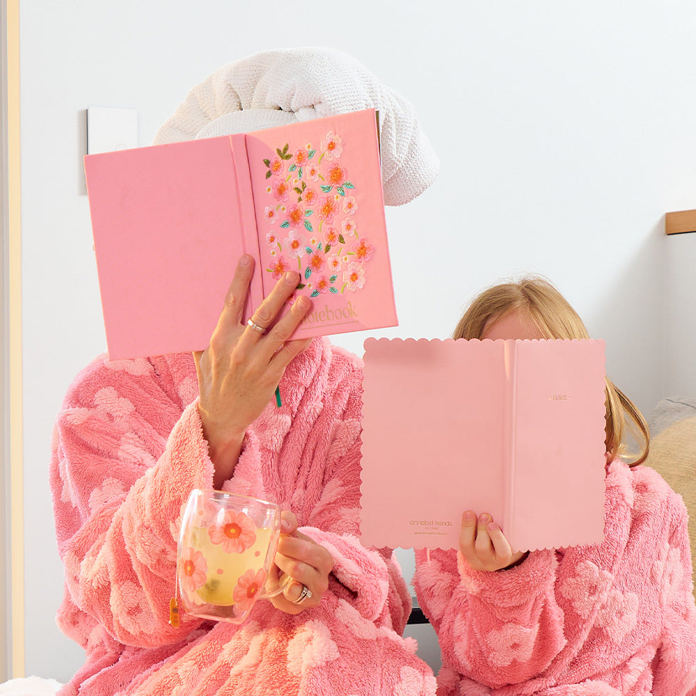 Two people in pink robes reading pink books with floral designs, holding drinks.