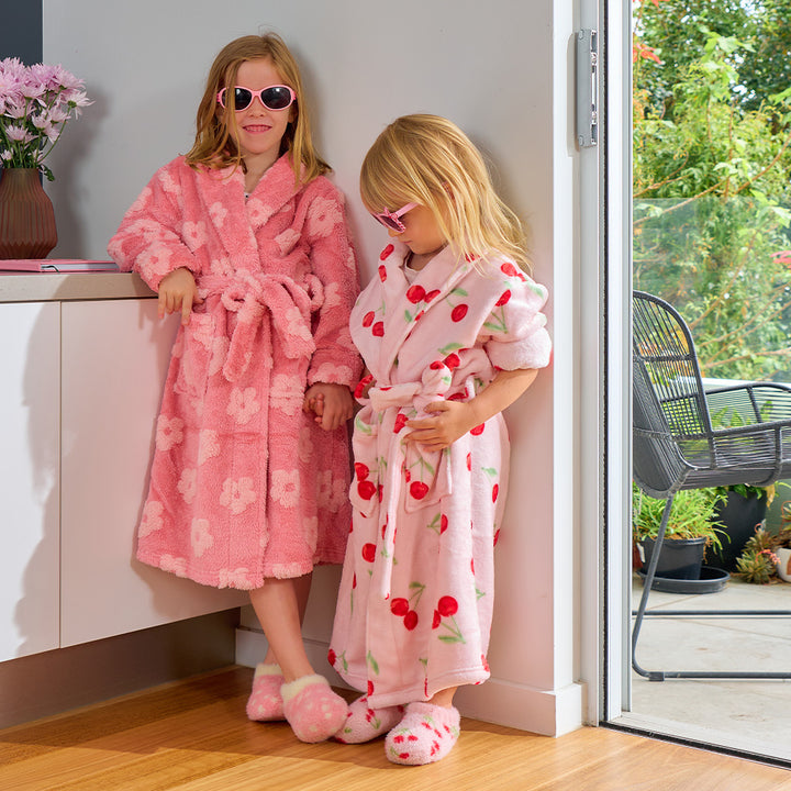 Two children in pink bathrobes standing in a room with a view of greenery outside.