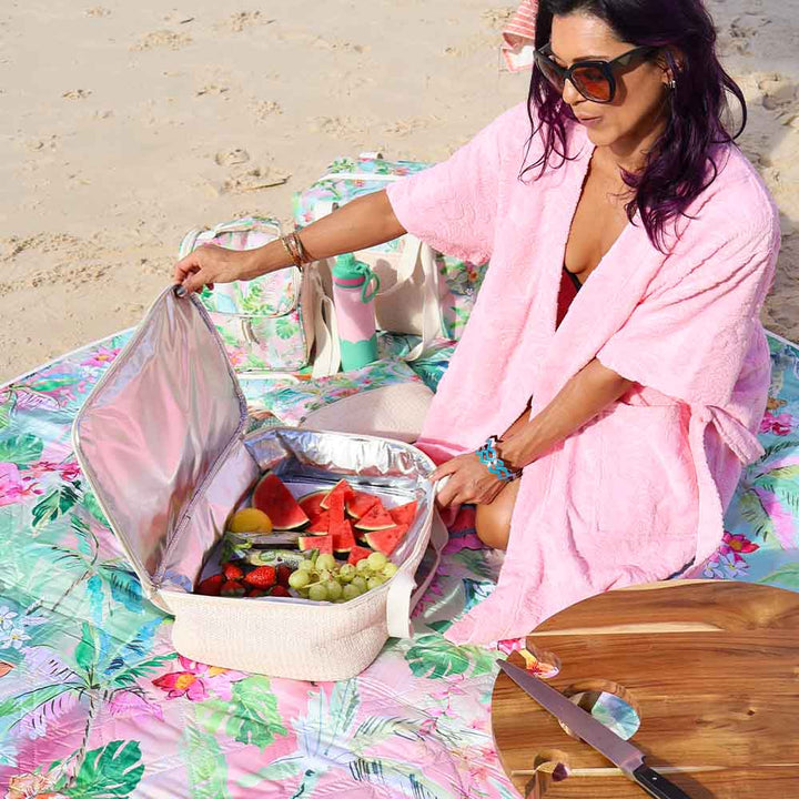 Woman in pink robe preparing a picnic on a beach with a colorful blanket and fruit.
