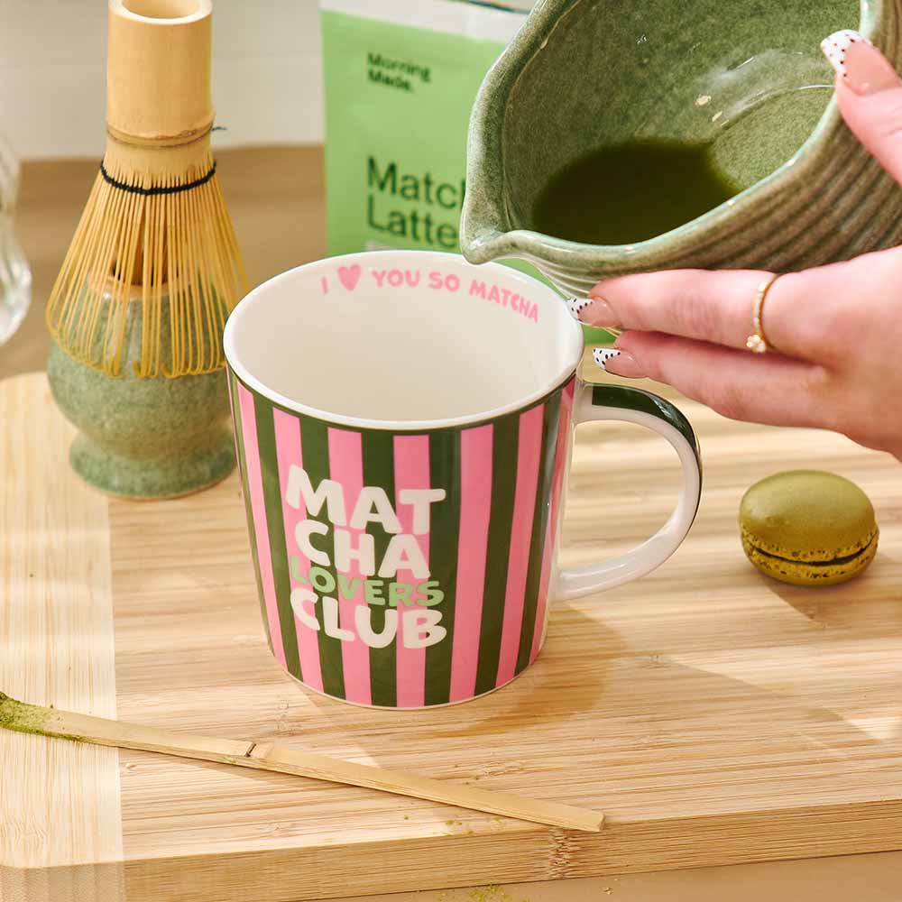Person preparing matcha latte with a striped mug labeled 'Matcha Lovers Club' on a wooden surface.