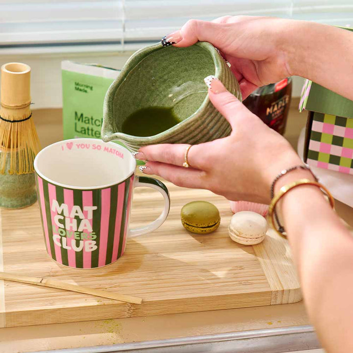 Person pouring matcha into a mug on a wooden surface with matcha-related items.