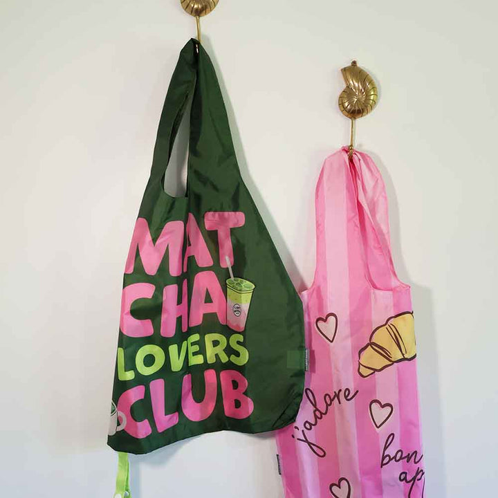 Two reusable bags, one green with pink and yellow text, the other pink with heart and croissant design, hanging on hooks against a white wall.