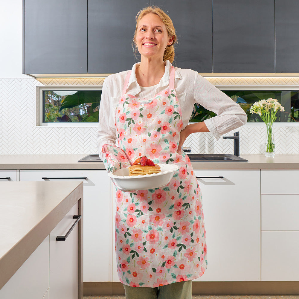 Woman in a kitchen wearing a floral apron holding a plate of pancakes.