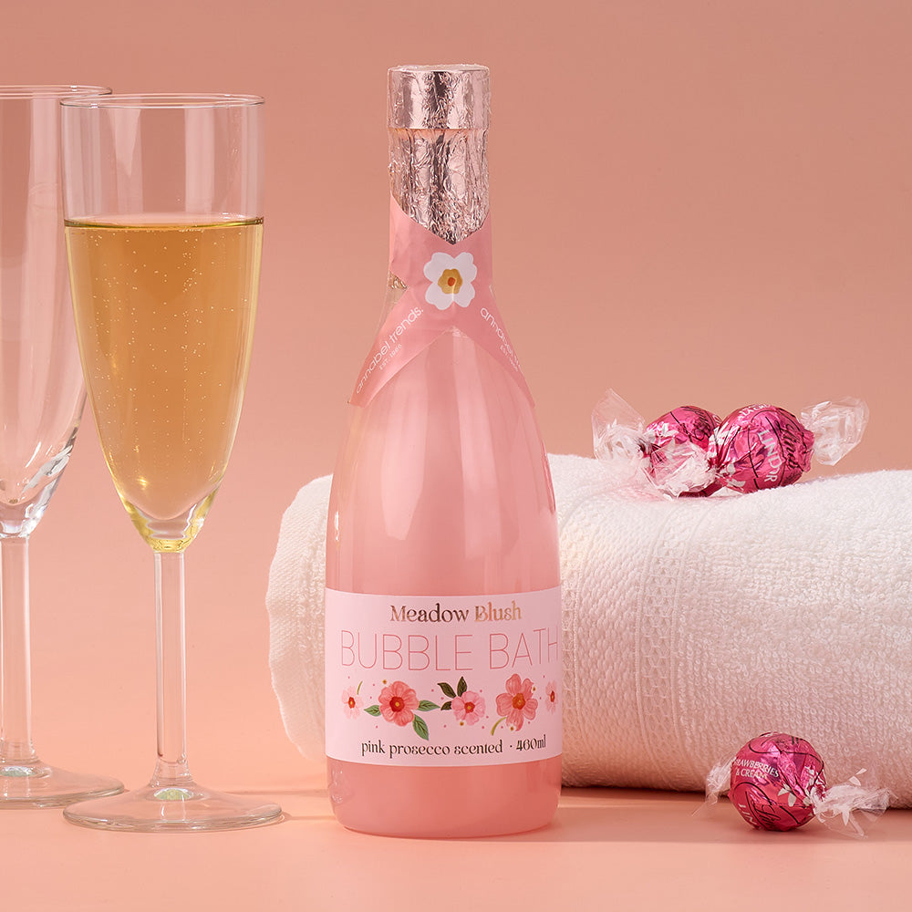 Pink bottle of 'Meadow Blush' bubble bath with a glass of champagne and wrapped candies on a pink background