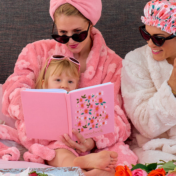 Woman and child in pink robes reading a pink notebook with floral embroidered design, surrounded by flowers.