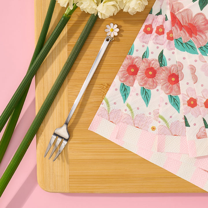 Floral-patterned napkin, fork, and flowers on a wooden board with a pink background