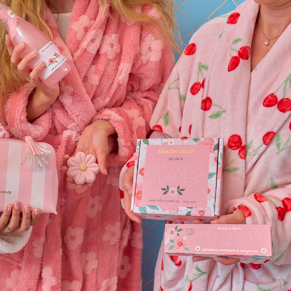 Two people in pink robes holding skincare products in meadow blush range  with a blue background